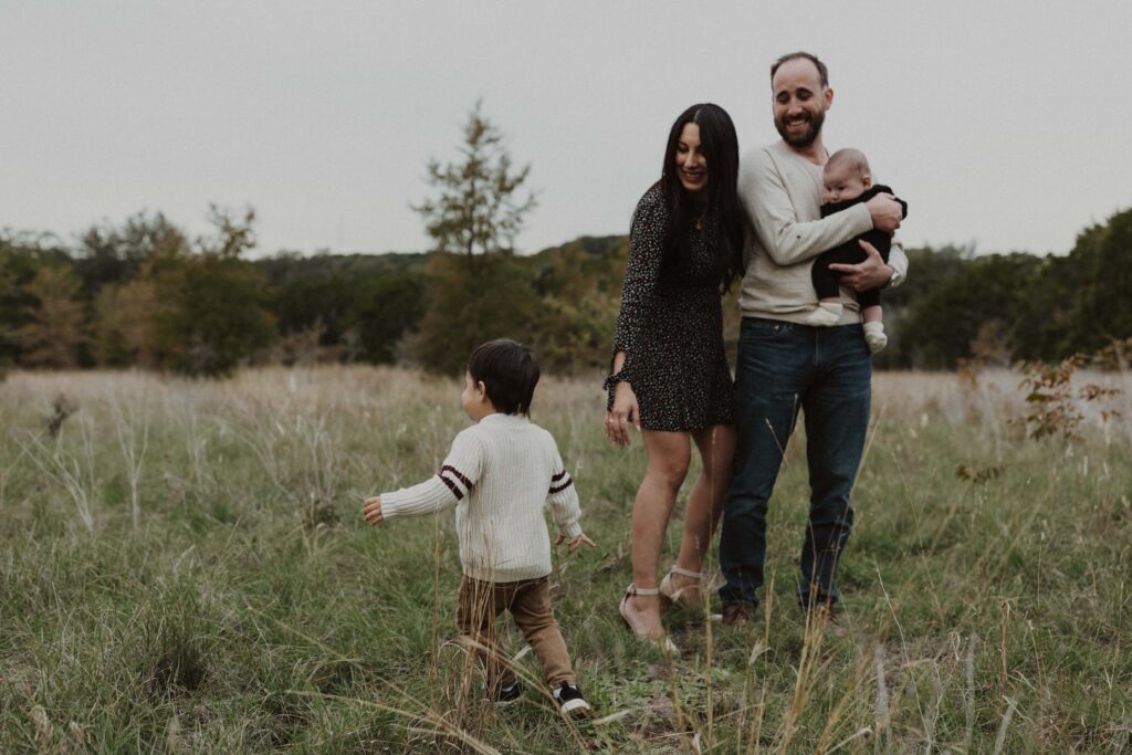 Mom and Dad laughing at their toddler running around in the grass during their Texas hill country family photos.