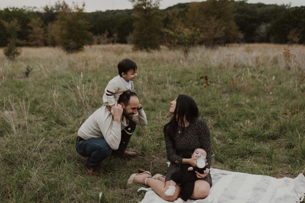 Mom feeding baby and Dad carrying toddler on his shoulders during their Texas hill country family photos.
